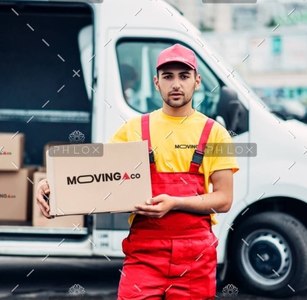 Delivery worker in a yellow shirt and red overalls holds a MOVING CO cardboard box in front of a white delivery van.