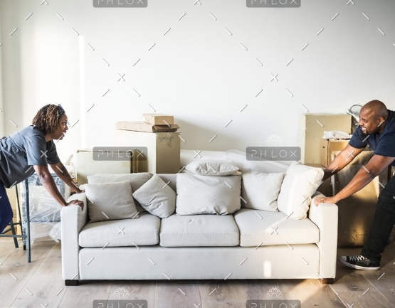 Two people push a white sofa into a bright living room with stacked moving boxes in the background.